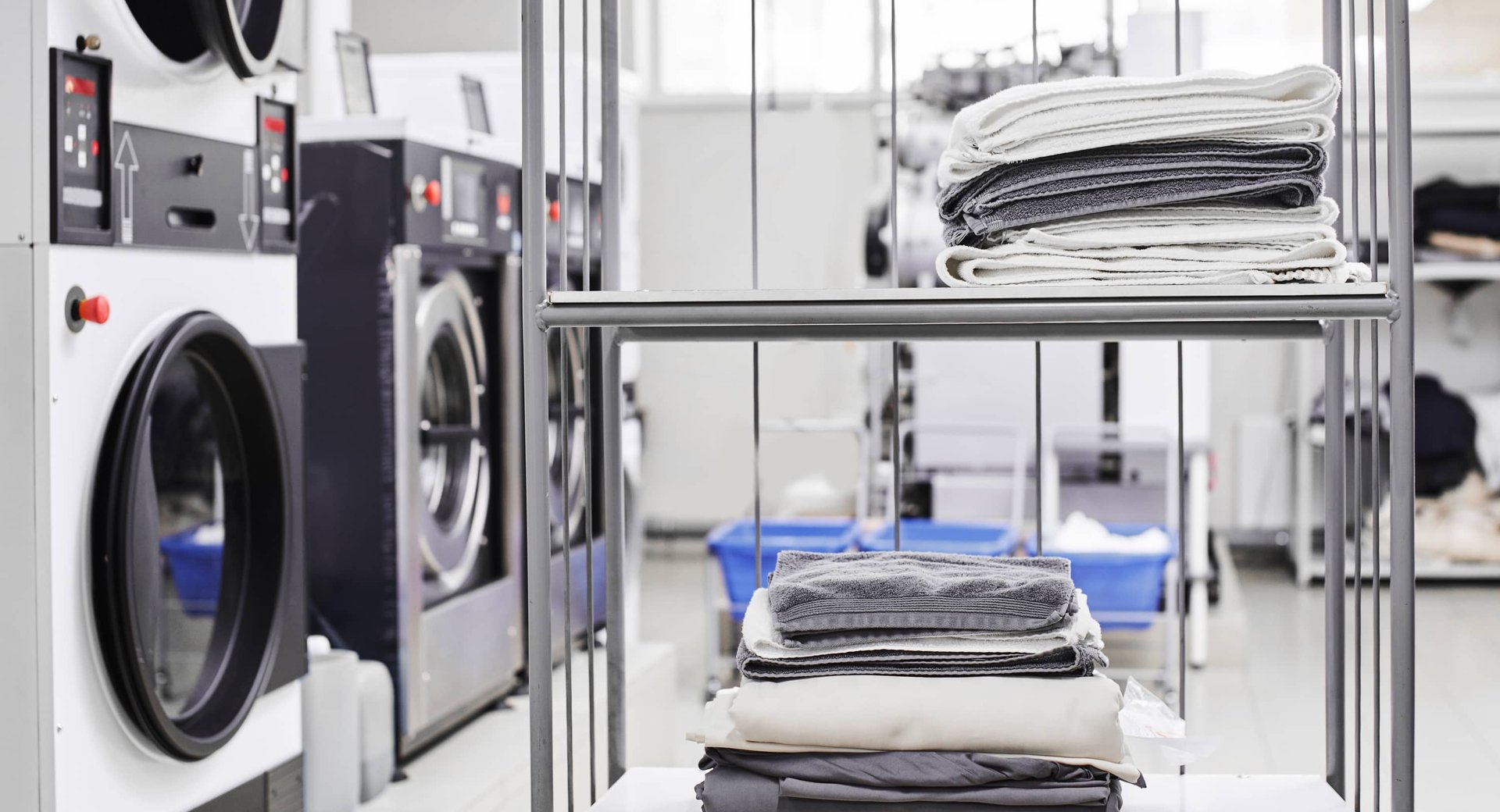 Metal cart with folded linens and towels beside commercial washers in laundromat